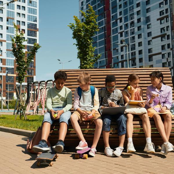 Photo by Norma Mortenson on pexels.com The image shows 5 children sitting on a bench in an urban environment. Some of them are eating, one girl is looking at the screen of her laptop.